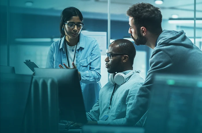 Three people collaborating in front of computer monitors in a modern office, with one person wearing headphones around the neck.
